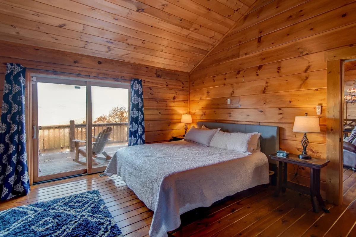 A serene bedroom featuring a comfortable bed positioned beside a sliding glass door, which opens out to a deck. The space is accented by a nightstand and a decorative rug. Natural light floods the room, highlighting the inviting atmosphere.