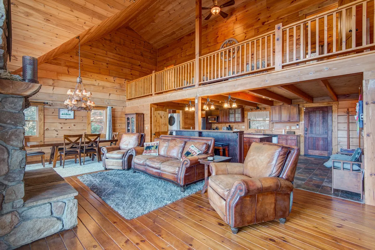 An inviting open-plan living area blending a plush leather seating arrangement, a robust stone fireplace, a wooden ceiling, and large windows looking out onto the mountains. The image also includes the dining area and a glimpse of the kitchen island, emphasizing togetherness and seamless flow.