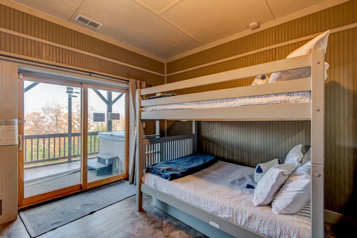 An inviting bunk bed area with a sliding glass door that opens onto a deck featuring a hot tub. This image showcases the seamless connection between indoor comfort and outdoor relaxation, with views of nature from the cozy interior and the promise of starlit evenings in the hot tub.