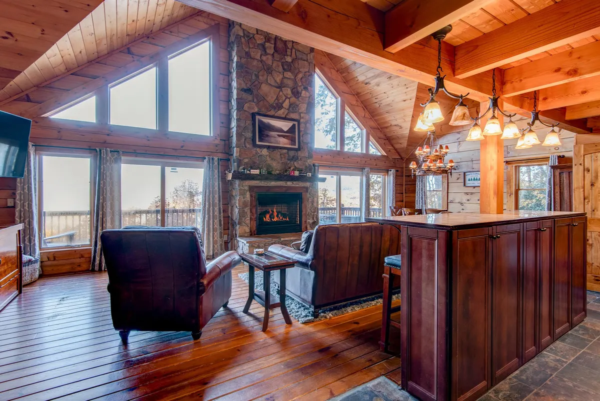 A grand great room featuring a large stone fireplace as its centerpiece, with vaulted wooden ceilings overhead and expansive windows that showcase sweeping mountain views. The comfortable seating area is perfect for gatherings or relaxing by the fire, and the open concept design hints at the adjacent kitchen and dining spaces.