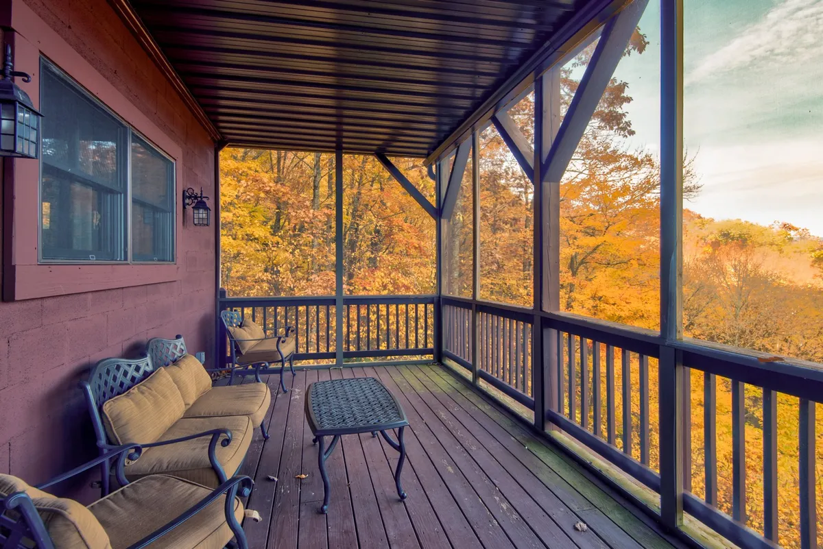 A screened porch surrounded by vibrant fall foliage with a comfortable seating arrangement, inviting guests to relax while immersed in nature. The porch is elevated, giving views of the trees and providing a safe, contained outdoor space that pets can enjoy alongside their owners.
