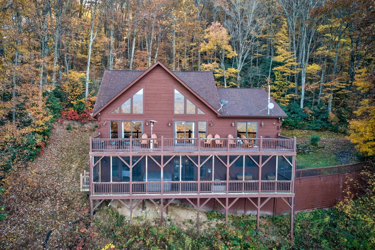A spacious wooden deck wraps around the exterior of the cabin, surrounded by vibrant autumn foliage. The deck features seating ideal for relaxing or socializing, with large windows offering a view inside. The inviting atmosphere suggests a pet-friendly space for guests and their dogs to roam or lounge while enjoying the mountain air.