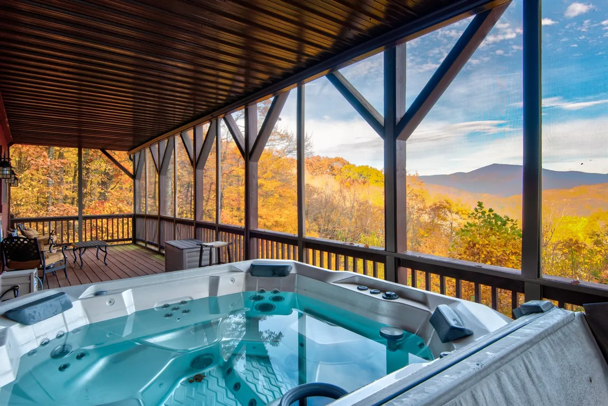The image showcases the cabin's inviting outdoor hot tub on the deck, overlooking expansive, layered mountain views. The tranquil setting is emphasized by the screened porch where guests can gather, with the golden hues of a mountain sunset painting the sky. The atmosphere feels serene and perfect for unwinding, either soaking in warmth or socializing with friends and family against a breathtaking natural backdrop.