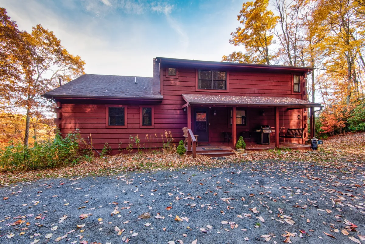 The front of the cabin features a wooden porch with Adirondack chairs, large windows, and surrounding fall foliage. The porch offers a welcoming entry point and a peaceful spot for coffee, thoughtfully designed for both owners and their pets to enjoy the fresh mountain air.