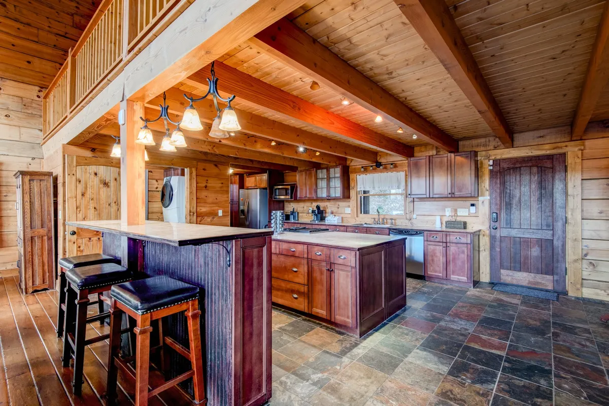 A modern kitchen centered around a granite-topped island with bar stools, accented by warm wooden beams and cabinetry. The space is designed for social cooking and casual dining, with stainless appliances visible in the background.