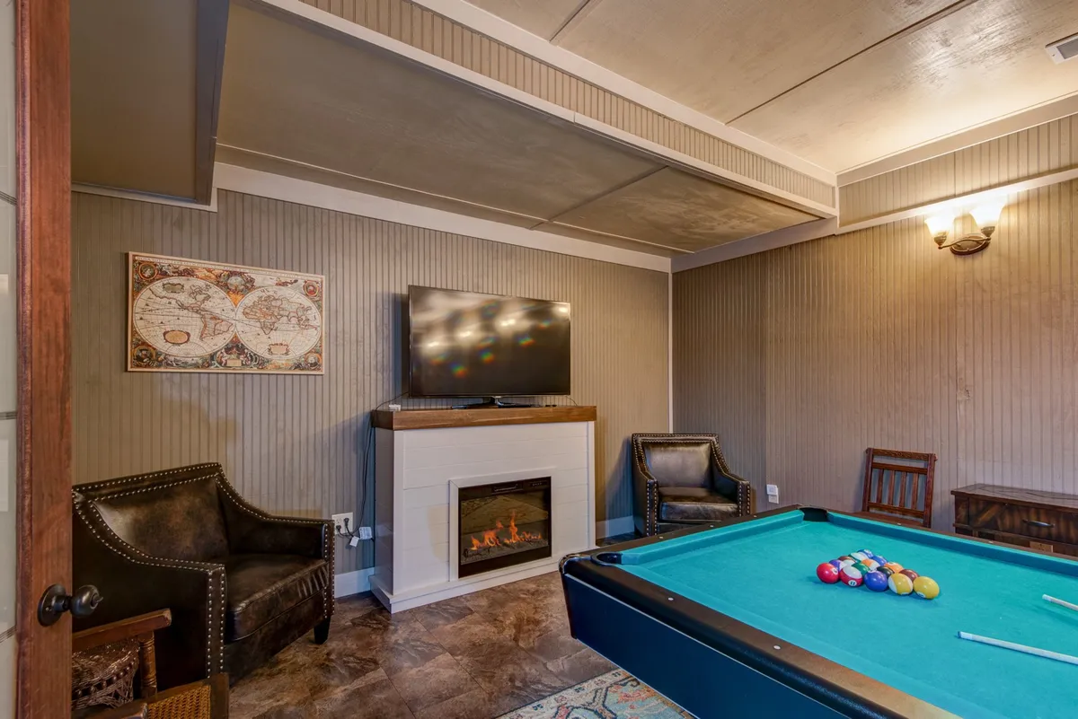 This inviting game room features a central pool table, a cozy fireplace, a large TV, and a comfortable seating area, creating the perfect hub for both friendly competition and movie nights. Warm wood finishes and soft lighting enhance the relaxed, entertaining vibe.